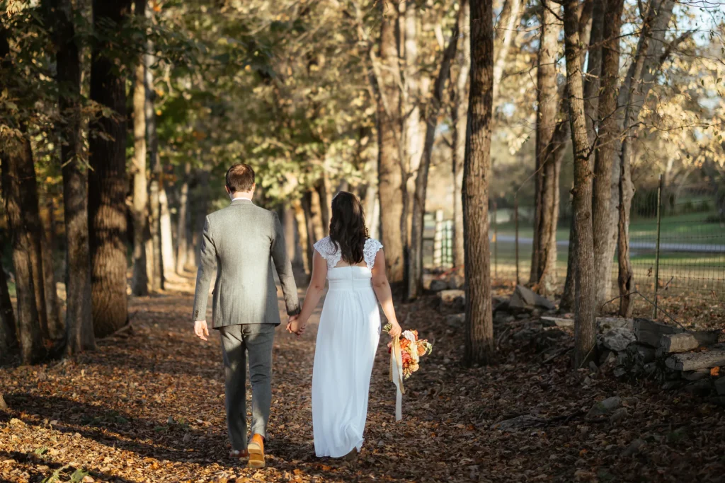 Newlyweds walking hand in hand down a leaf-covered woodland path in autumn sunlight.
