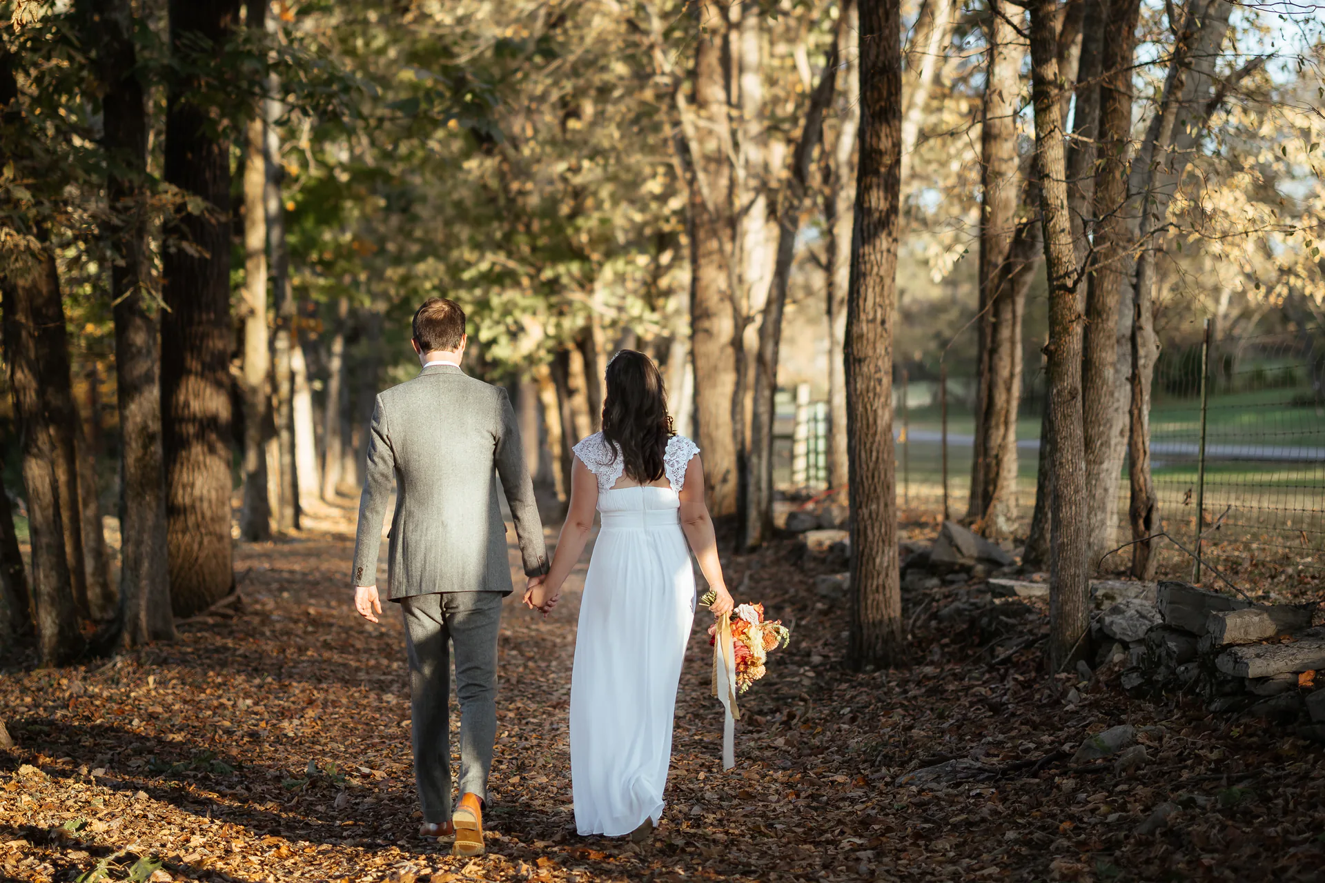Newlyweds walking hand in hand down a leaf-covered woodland path in autumn sunlight.