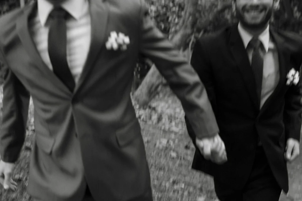 Two grooms in suits with boutonnieres holding hands as they walk together outdoors