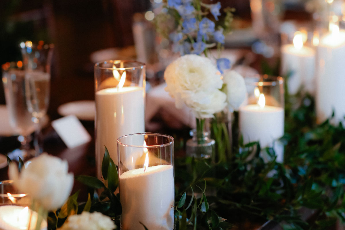 Pillar candles in glass hurricanes along a table runner of greenery, white ranunculus and blue delphinium