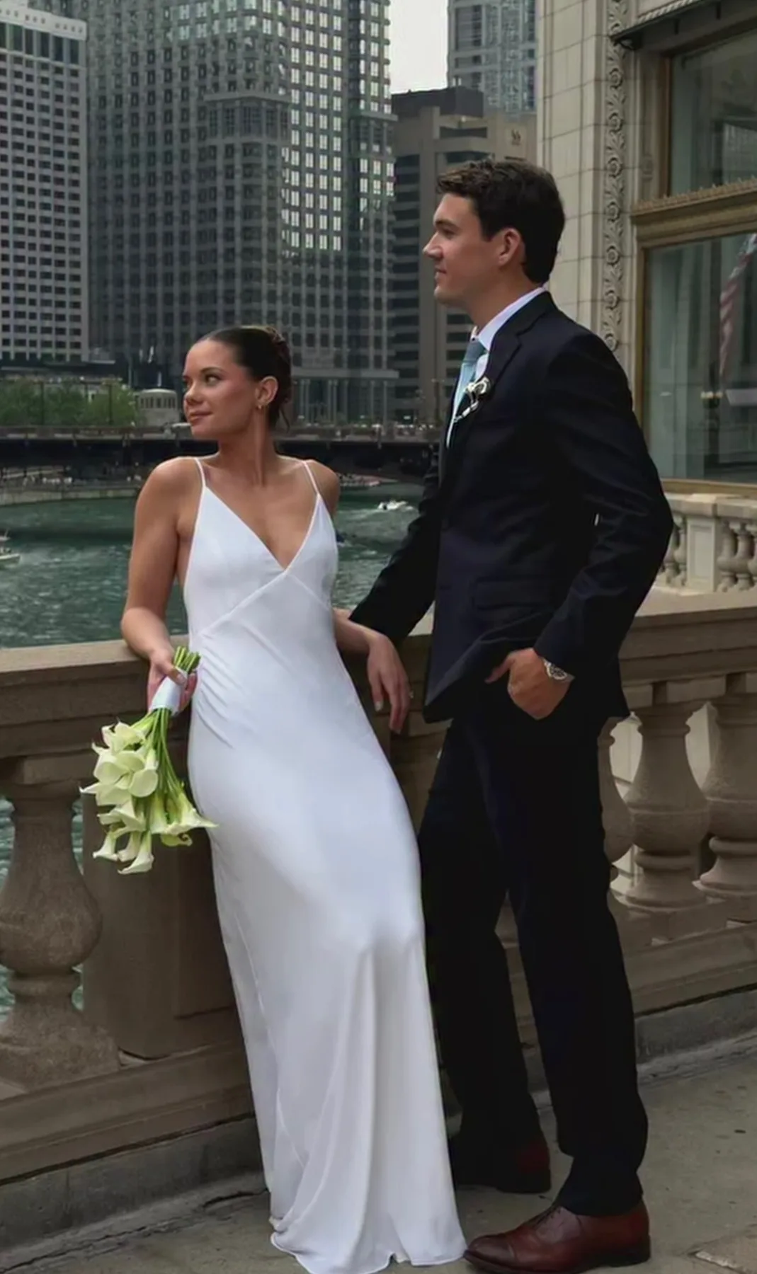 Bride in a white gown holding a bouquet of white calla lilies beside a groom in a dark suit along a riverfront railing with city buildings in the background.