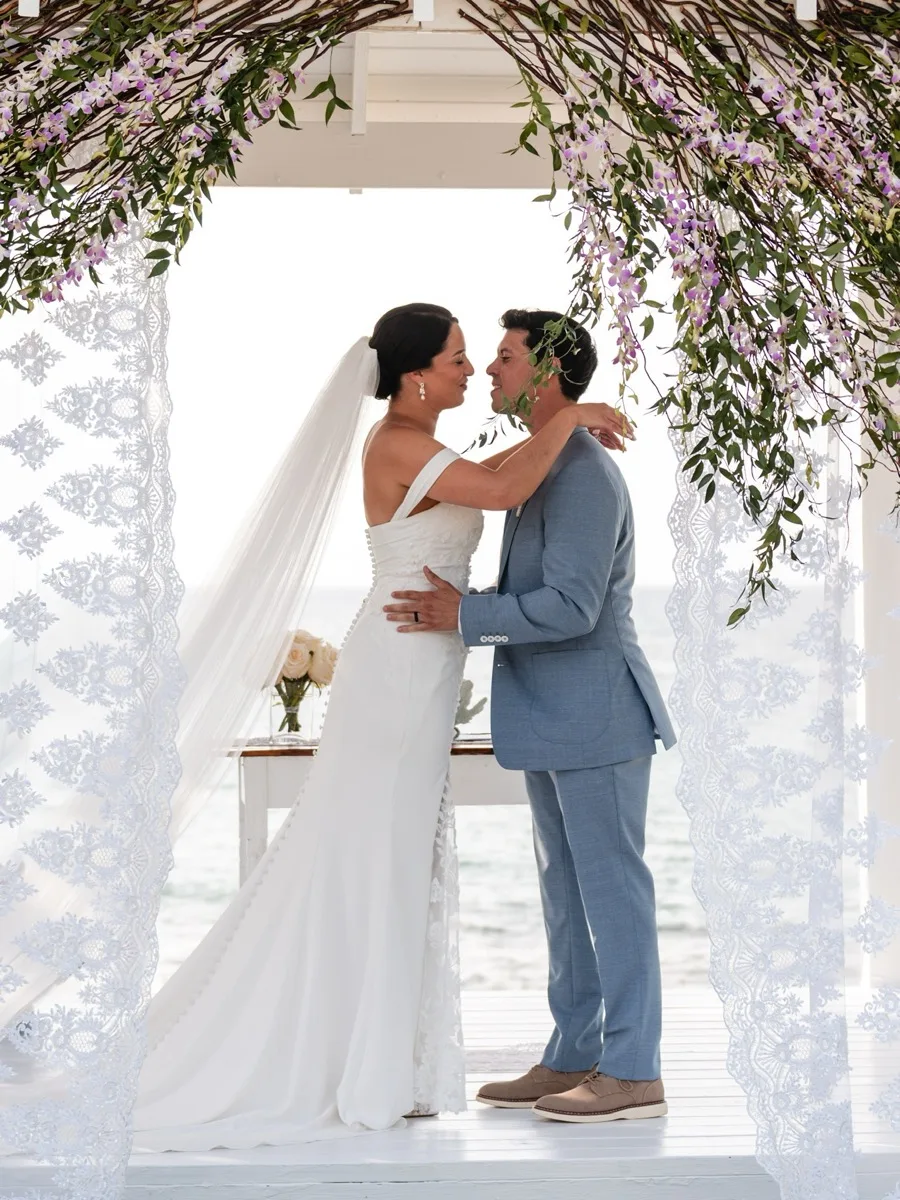 Bride and groom about to kiss beneath an orchid and lace draped seaside pavilion, the ocean behind them