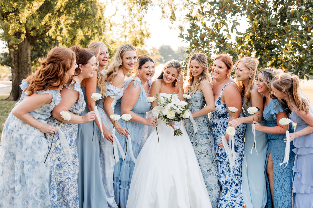 Bride laughing with her bridesmaids in blue and periwinkle gowns, each holding a single white rose, under trees in golden evening light