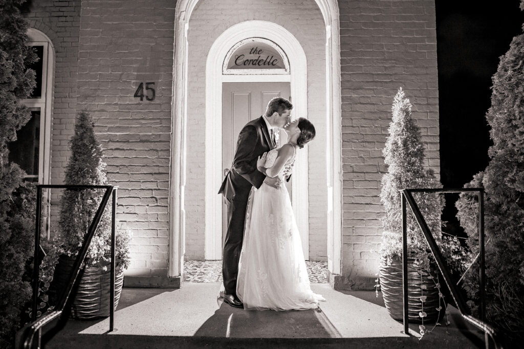 Black and white photo of newlyweds kissing at a brick wedding venue entrance
