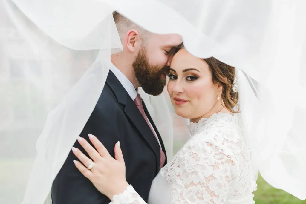 Close-up portrait of a bride in a high-neck lace wedding gown and a bearded groom in a dark suit embraced beneath a flowing white veil, with the bride's hand resting on his chest showing her wedding ring.