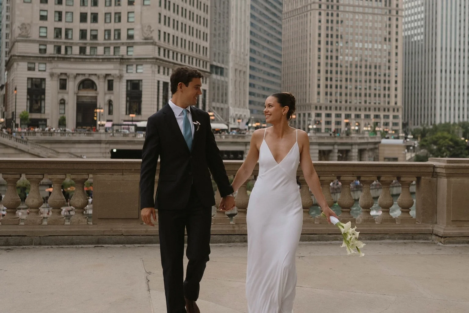 Bride and groom holding hands as they walk beside a stone balustrade above the Chicago River, with the downtown skyline behind them